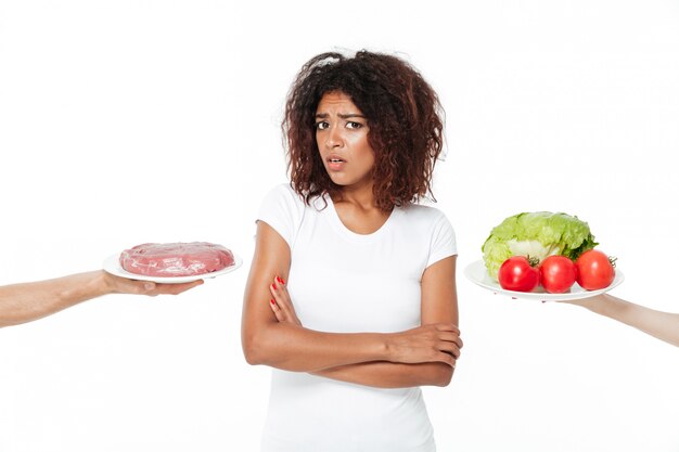 Mujer africana joven confusa que elige entre la carne y las verduras.