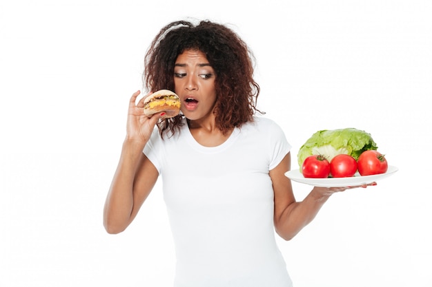 Mujer africana joven confusa que elige entre la hamburguesa y las verduras.