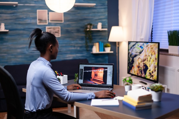 Mujer africana joven que prueba el juego profesional en línea en la computadora portátil en casa a altas horas de la noche. Jugador profesional que controla los videojuegos digitales en su computadora con tecnología inalámbrica de red moderna.