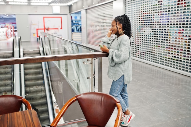 Mujer africana posada en suéter y jeans posada en el centro comercial sentada en la mesa con teléfono móvil