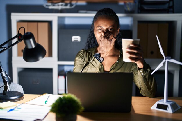 Mujer africana que trabaja usando una computadora portátil por la noche oliendo algo apestoso y repugnante olor intolerable aguantando la respiración con los dedos en la nariz mal olor