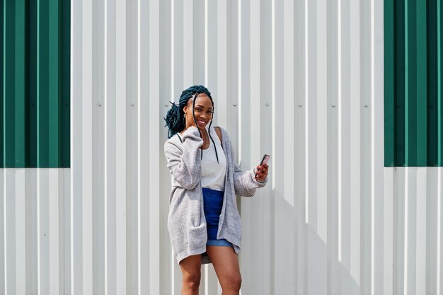 Mujer africana con rastas en pantalones cortos de jeans posada contra una pared de acero blanco con teléfono móvil en la mano