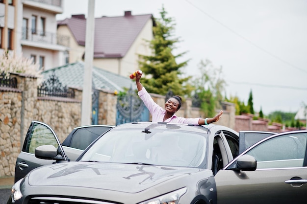 Mujer africana de ricos negocios en coche suv plateado en techo corredizo divirtiéndose con cerveza a mano