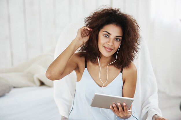 Mujer africana en ropa de dormir sonriendo escuchando música en auriculares sentado en la cama.