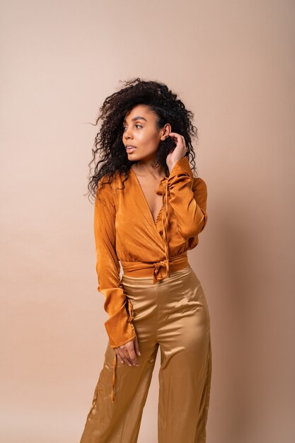 Mujer africana segura con cabellos rizados perfectos en blusa naranja casual y pantalón dorado posando en la pared beige.