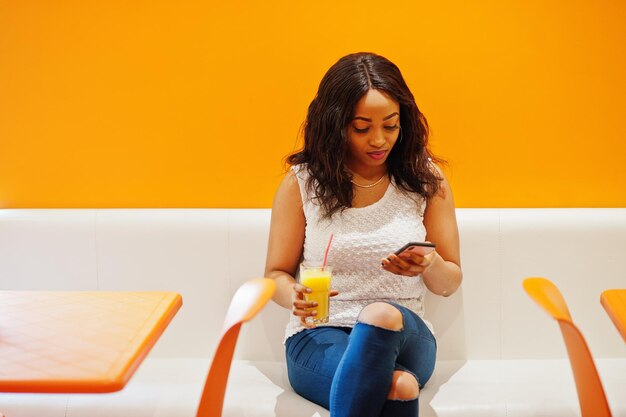 Mujer africana sentada en un café contra la pared naranja con jugo de piña y teléfono móvil en las manos