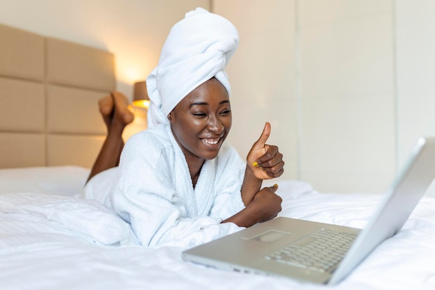 Mujer africana sonriente acostada en la cama en bata de baño con una computadora portátil hablando con sus amigos a través de una videollamada Mujer africana relajándose en la cama después del baño y mirando su computadora portátil