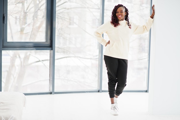 Mujer africana en sudadera con capucha stand apartamento interior cerca de grandes ventanales