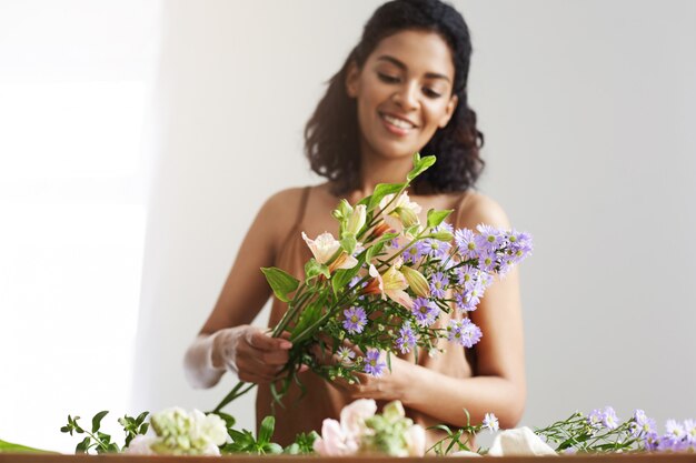 Mujer africana tierna feliz que sonríe haciendo el ramo de flores en el lugar de trabajo sobre la pared blanca.