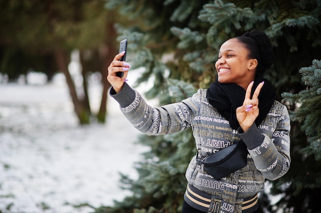 Mujer africana vestida con bufanda negra parada cerca del árbol de año nuevo en el día de invierno en Europa con teléfono móvil en las manos y haciendo selfie