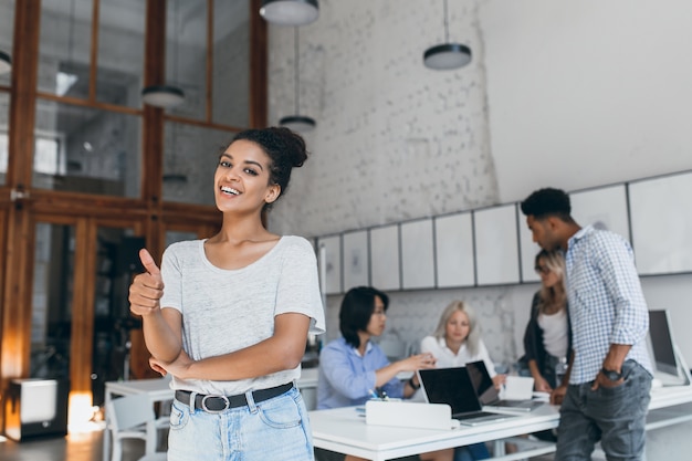 Mujer africana viste jeans azul claro y cinturón negro disfrutando del trabajo en equipo con colegas internacionales. Especialistas independientes mujeres con estilo posando mientras sus amigos trabajan con la computadora portátil.