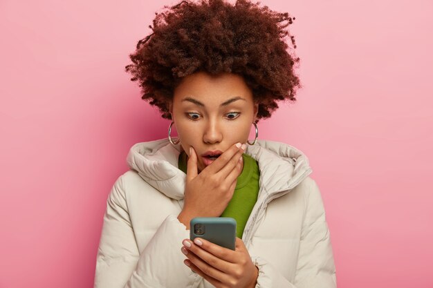 Mujer afro aturdida toca la barbilla por la conmoción, tiene el pelo negro rizado, viste bata blanca, posa contra la pared rosa del estudio