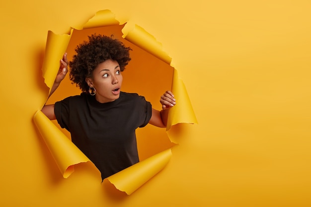 Mujer afro en camiseta negra jadea de asombro, mira con expresión asustada a un lado, vestida con camiseta negra