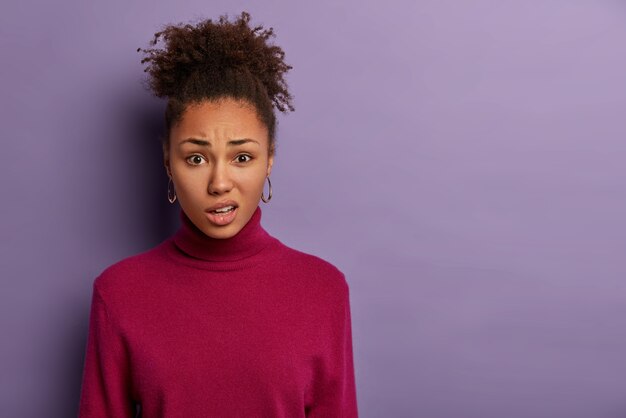 Mujer afro disgustada sonríe y frunce los labios, reacciona al escuchar algo desagradable