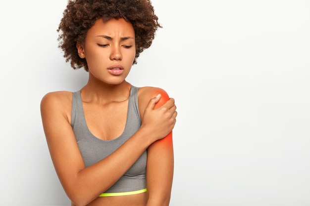 Mujer afro insatisfecha toca el hombro rojo, estira los músculos durante el entrenamiento deportivo, tiene expresión triste, usa sujetador gris