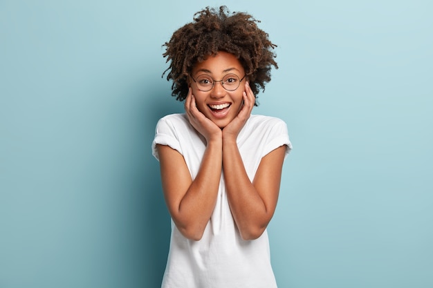 Mujer afro optimista despreocupada toca ambas mejillas, tiene una amplia sonrisa, muestra los dientes blancos, vestida con ropa informal, gafas redondas
