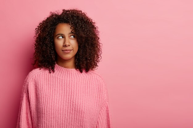 La mujer afro piensa profundamente en algo, piensa cómo actuar en una situación difícil, viste un suéter de punto rosa en el interior