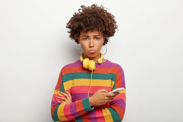 Mujer afro rizada molesta con mirada triste, usa teléfono móvil, no puede retirar dinero de una cuenta en línea, usa auriculares para escuchar música