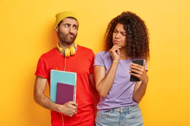 Una mujer afro seria sostiene la barbilla, usa un teléfono inteligente moderno, mira sospechosamente a su novio, verifica que su casilla de correo electrónico está celosa. Estudiante masculino con Bloc de notas