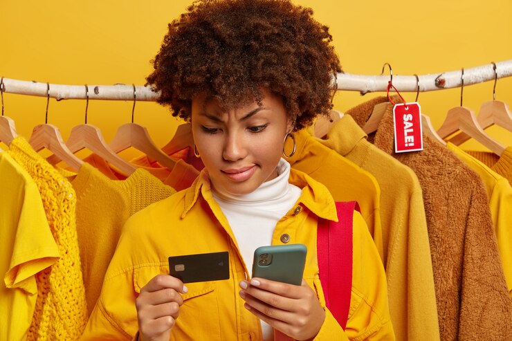 Foto gratuito mujer afro seria usa tarjeta de crédito con teléfono móvil para compras en línea en grandes almacenes, compra ropa en oferta, vestida con camisa amarilla de moda, se opone a diferentes prendas en perchas
