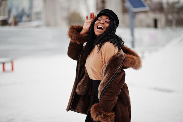Mujer afroamericana con abrigo de piel de oveja y gorra posada en el día de invierno contra un fondo nevado