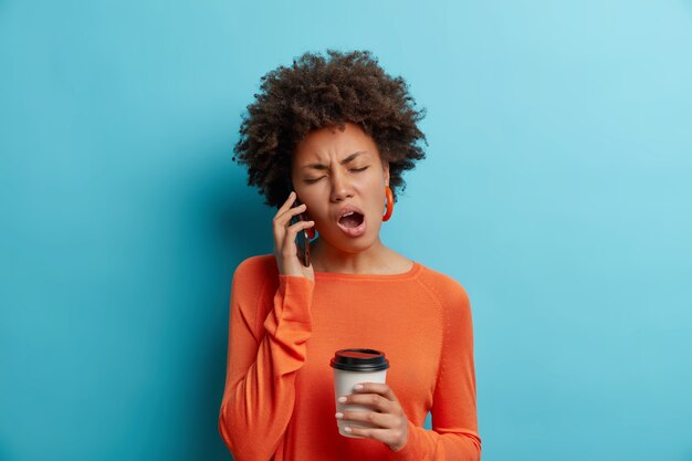 Mujer afroamericana aburrida y soñolienta escucha una historia poco interesante mientras habla a través de un teléfono inteligente, bebidas, café para llevar, usa un jersey naranja con aretes aislados sobre una pared azul