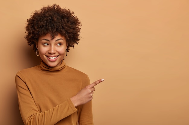 La mujer afroamericana alegre y complacida indica a un lado, promociona productos, sonríe con alegría, usa un jersey de cuello alto marrón, posa contra una pared marrón. Concepto de publicidad. Cosas bonitas esto. Mira ahí