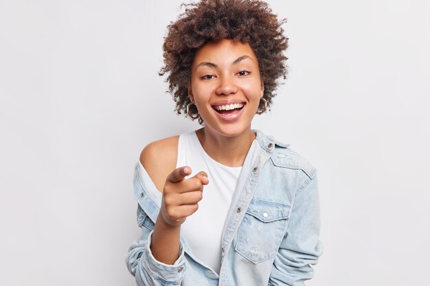 La mujer afroamericana alegre despreocupada apunta a que sonríe ampliamente ve algo divertido en el frente vestida con ropa de moda aislada sobre la pared blanca