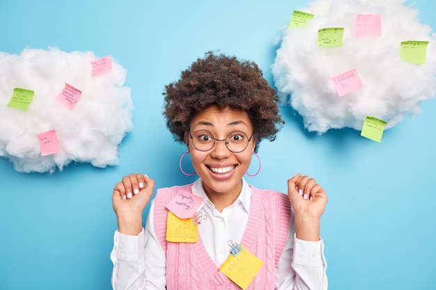 La mujer afroamericana alegre positiva levanta la mano mira con entusiasmo al frente, contenta de escuchar elogios por el buen trabajo, usa anteojos redondos, ropa formal rodeada de notas adhesivas con tareas escritas.