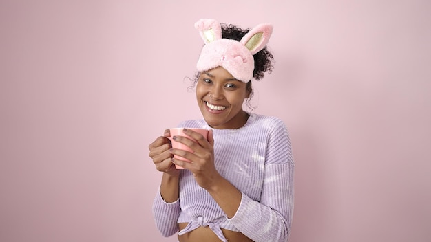 Mujer afroamericana con antifaz para dormir bebiendo una taza de café sobre un fondo rosa aislado