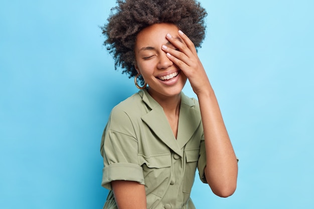 La mujer afroamericana bastante alegre mantiene la palma en la cara se siente muy contenta cierra los ojos sonríe ampliamente viste un vestido elegante aislado sobre una pared azul