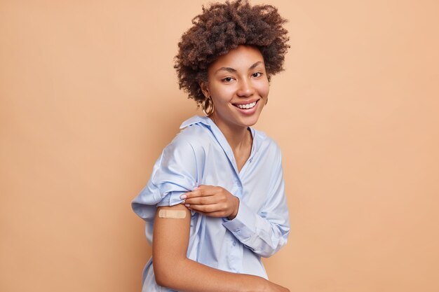 Mujer afroamericana bastante complacida en camisa azul levanta manga muestra brazo vacunado usa yeso adhesivo sonríe agradablemente aislado sobre pared beige