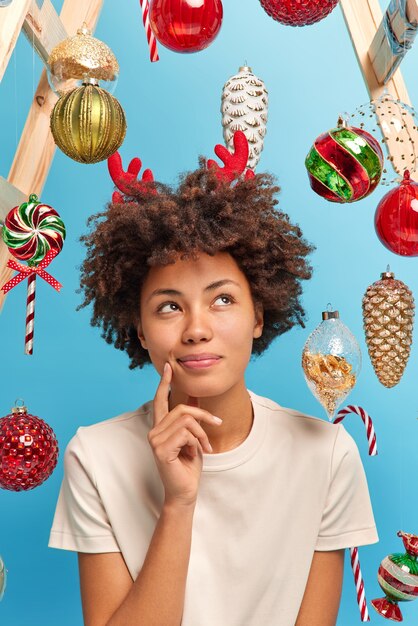 Una mujer afroamericana bastante pensativa posa pensativamente en el interior vestida con ropa casual y se ve arriba hace ideas para la celebración perfecta del Año Nuevo piensa en los regalos de Navidad para los familiares