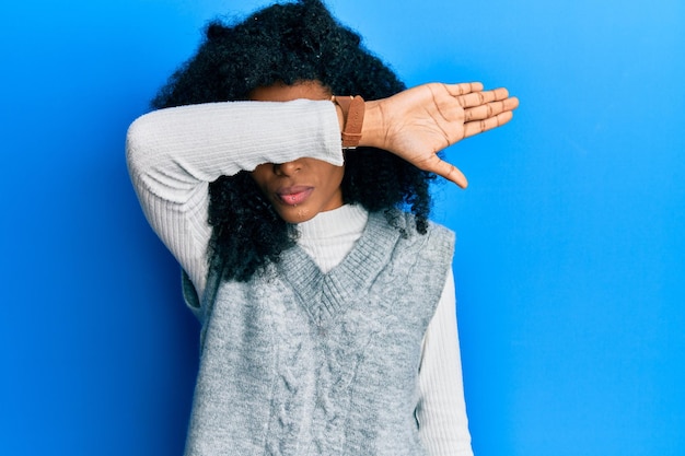 Mujer afroamericana con cabello afro que usa un suéter de invierno casual que cubre los ojos con el brazo que parece serio y triste concepto de escondite y rechazo sin vista