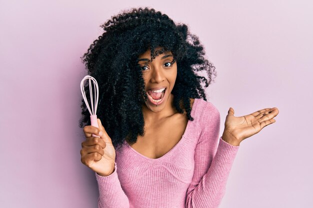 Mujer afroamericana con cabello afro sosteniendo un batidor pequeño celebrando el logro con una sonrisa feliz y expresión ganadora con la mano levantada