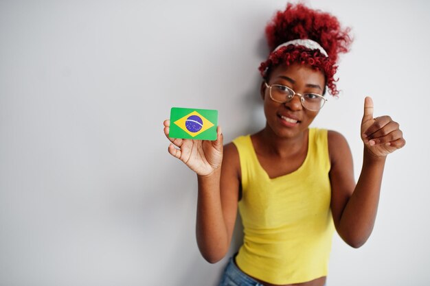 Mujer afroamericana con cabello afro usa camiseta amarilla y anteojos sostiene la bandera de Brasil aislada sobre fondo blanco muestra el pulgar hacia arriba