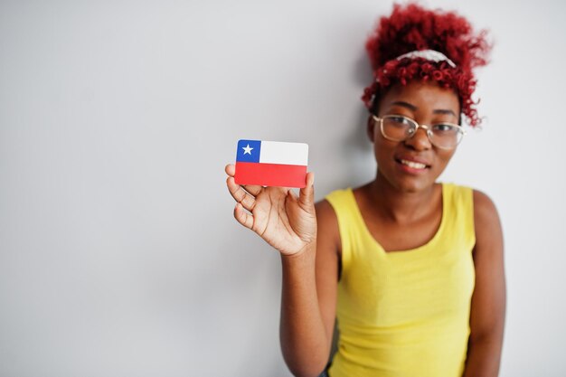 Mujer afroamericana con cabello afro usa camiseta amarilla y anteojos sostiene la bandera de Chile aislada en fondo blanco