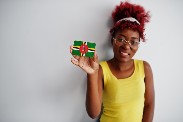 La mujer afroamericana con cabello afro usa una camiseta amarilla y anteojos sostiene la bandera de Dominica aislada en el fondo blanco