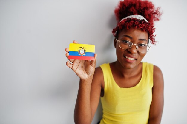 Mujer afroamericana con cabello afro usa camiseta amarilla y anteojos sostiene la bandera de Ecuador aislada sobre fondo blanco