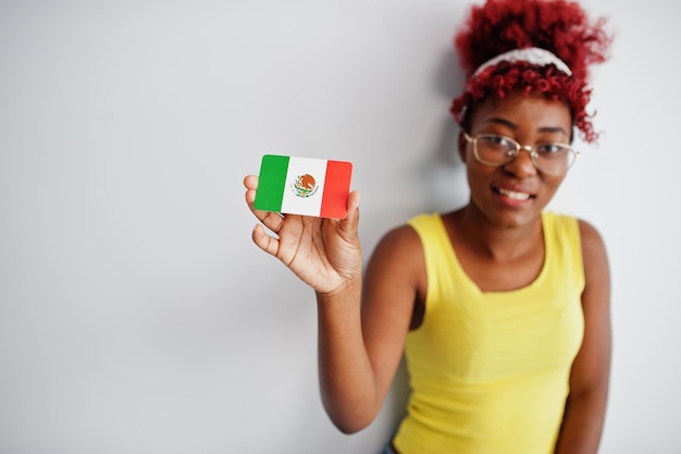 Mujer afroamericana con cabello afro usa camiseta amarilla y anteojos sostiene la bandera de méxico aislada en fondo blanco