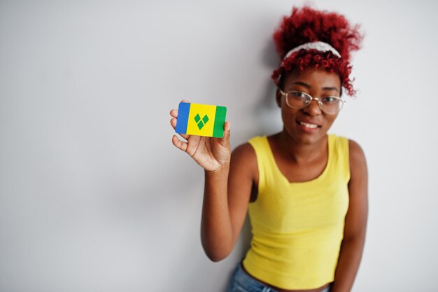 Mujer afroamericana con cabello afro usa camiseta amarilla y anteojos sostiene la bandera de San Vicente y las Granadinas aislada sobre fondo blanco