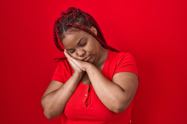 Mujer afroamericana con cabello trenzado sobre fondo rojo durmiendo cansada soñando y posando con las manos juntas mientras sonríe con los ojos cerrados.