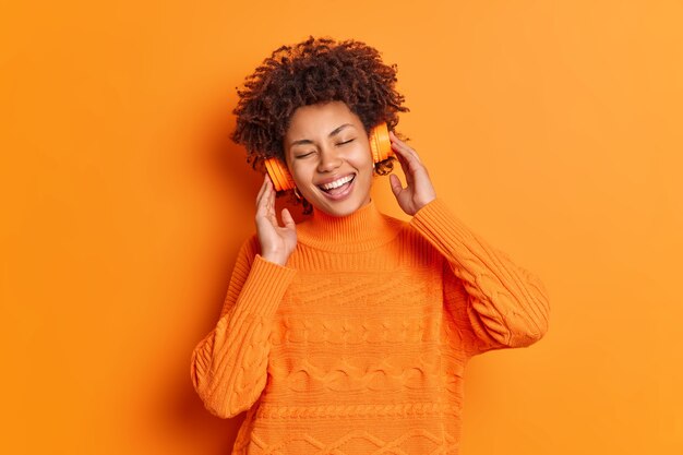 Mujer afroamericana complacida disfruta de una canción agradable a través de auriculares estéreo sonríe ampliamente mantiene los ojos cerrados tiene expresión feliz