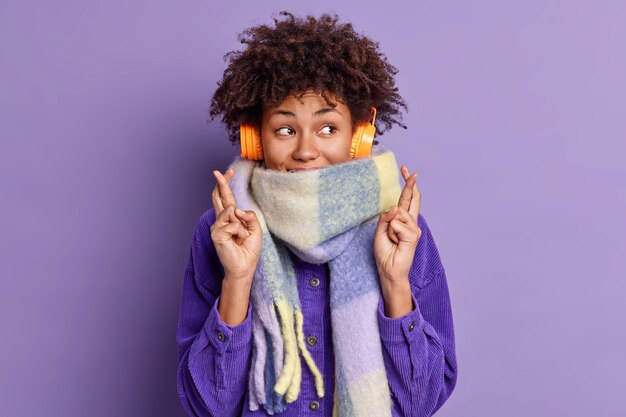 La mujer afroamericana complacida de pelo rizado cruza los dedos desea lo mejor, cree en la buena fortuna, usa una bufanda cálida de invierno alrededor del cuello, escucha su música favorita a través de auriculares