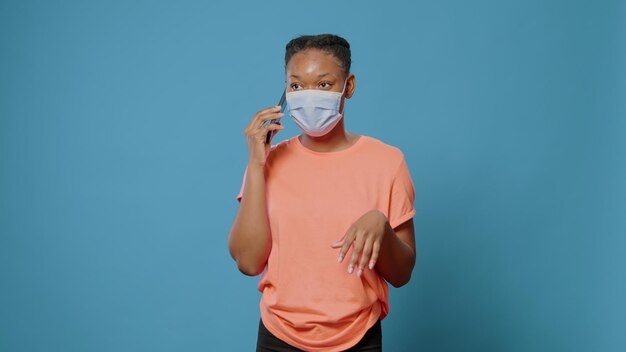 Mujer afroamericana conversando por teléfono mientras usa mascarilla contra el coronavirus. Persona joven que usa un teléfono inteligente para hablar con amigos en el estudio durante la pandemia de covid 19.