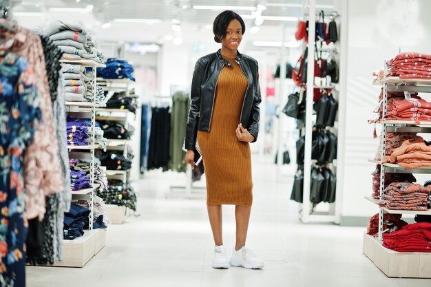 Mujer afroamericana delgada con vestido de túnica marrón y chaqueta de cuero negro posada en la tienda de ropa Es hora de ir de compras