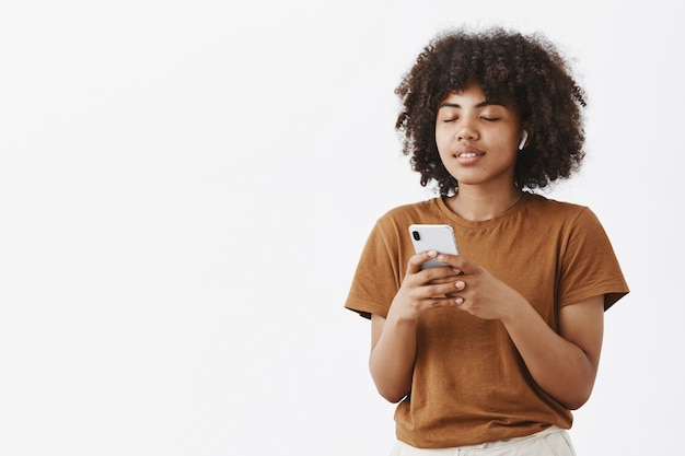 Mujer afroamericana despreocupada complacida tranquila y relajada en camiseta marrón cerrando los ojos sosteniendo el teléfono inteligente usando auriculares inalámbricos