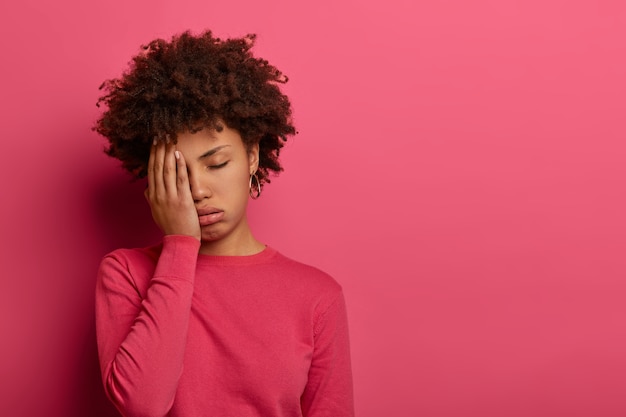 Mujer afroamericana disgustada cubre la cara con la palma, se siente muy cansada y agotada, no puede seguir trabajando