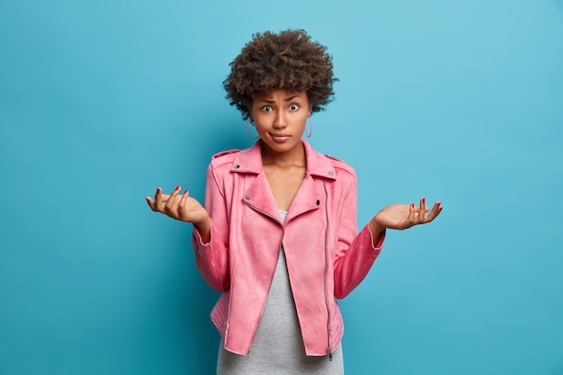 Mujer afroamericana dudosa insegura con cabello rizado extiende las palmas de las manos hacia los lados, molesta por hacer una elección, vestida con una chaqueta rosa de moda, se queda descuidada, no puede responder a la pregunta, posa en el interior