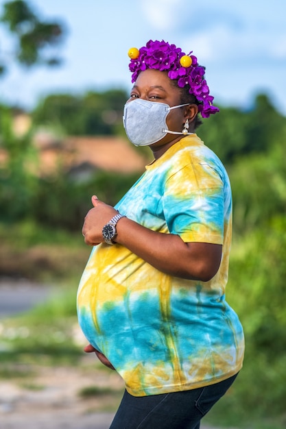 Mujer afroamericana embarazada en una mascarilla protectora gesticulando el signo de los pulgares hacia arriba